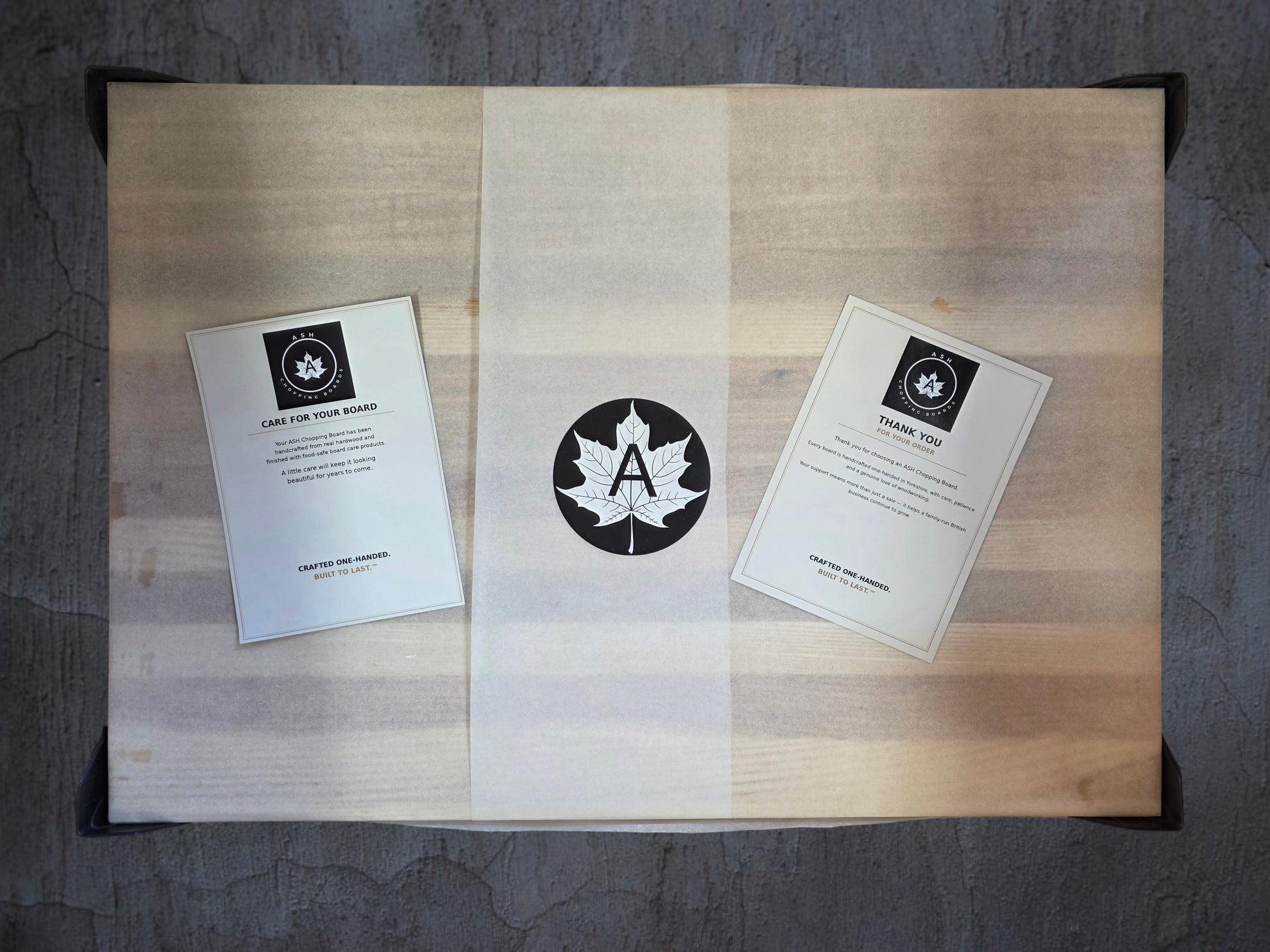 A custom wooden gift box with a light-colored wood grain finish, sealed in clear plastic packaging. Inside, a white paper sleeve with a black circular maple leaf logo containing the letter 'A' is wrapped around the center, flanked by two white cards with black text and logos.